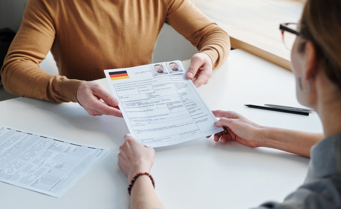 Unrecognizable man sitting in front of visa agency specialist giving application form papers with photos to her